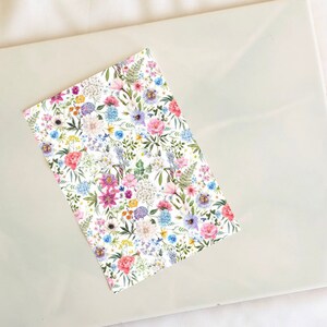 Pode incluir: Um fundo branco com um padrão floral com flores rosa, azuis, amarelas e roxas. As flores são cercadas por folhas verdes.
