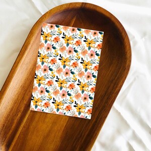May include: A white card with a floral pattern featuring yellow, orange, and pink flowers on a white background. The card is sitting on a wooden tray.