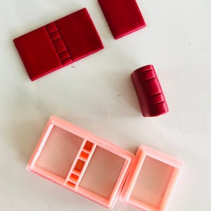 Könnte beinhalten: Ein Satz aus vier roten Tonstücken zum Herstellen eines Miniaturbuches. Die Stücke haben die Form eines Buchdeckels, eines Rückens und zweier Seiten. Außerdem gibt es zwei rosa Plastikformen zum Herstellen des Buches.