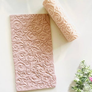 May include: A pink rolling pin with a decorative swirl pattern. The rolling pin is next to a flat piece of pink fondant with the same swirl pattern.