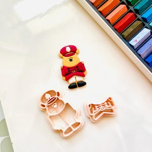 May include: A set of three cookie cutters, one shaped like a bear wearing a red and gold uniform, one shaped like a bear head, and one shaped like a bow. The cutters are made of white plastic.