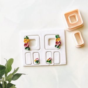 May include: A white polymer clay earring mold with square and rectangular cutouts, adorned with colorful floral accents. Two matching cutters are also present. The flowers feature yellow, pink, and green hues, suggesting a spring or summer theme.