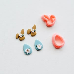 May include: Two pairs of polymer clay earrings, one pair is brown bunny shaped with white flowers and the other pair is blue teardrop shaped with white flowers. There are also two pink polymer clay cutters, one is a bunny shape and the other is a teardrop shape.