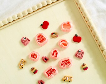 May include: A collection of pink and red plastic cookie cutters shaped like school supplies, including a bus, apple, backpack, pencil, and crayon.
