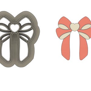 May include: A cookie cutter in the shape of a bow with a heart in the center. The cutter is made of metal and has a silver finish. The bow is tied with a ribbon and has a heart in the center.
