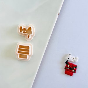 May include: Two white plastic cookie cutters shaped like a dog and a doghouse. A red and white fondant dog and doghouse are shown on a white surface.