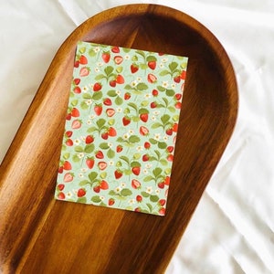 May include: A green card with a repeating pattern of red strawberries and white flowers. The card is sitting on a wooden tray.
