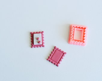Può includere: Tre tagliapasta di plastica rosa a forma di quadrato con bordi a festoni. I tagliapasta sono su una superficie bianca.