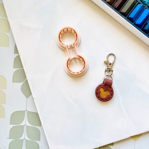 May include: A white surface with a pink plastic cookie cutter shaped like a figure eight with two circles. A brown leather keychain with a gold Mickey Mouse head charm is on the surface.