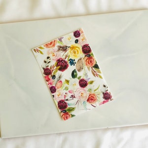 May include: A white card with a floral pattern featuring burgundy, pink, yellow, and green watercolor flowers. The flowers are arranged in a bouquet with feathers and leaves.