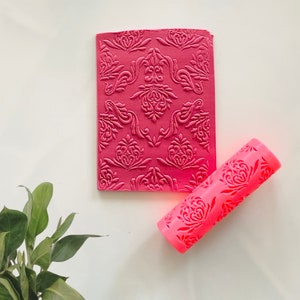 May include: A pink rolling pin with a decorative pattern and a pink silicone mat with a matching pattern. The mat is embossed with a damask design.