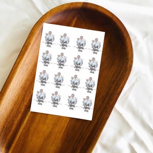 May include: A sheet of stickers with the text "Best Mom ever" in a script font, surrounded by a floral design. The stickers are on a wooden tray with a light-colored background.