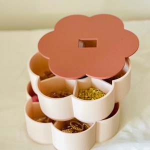 K&ouml;nnte beinhalten: Ein dreistufiger Schmuck-Organizer in Blumenform. Die Oberseite und der Deckel sind in einem ged&auml;mpften Rosaton gehalten, w&auml;hrend die F&auml;cher hellbeige sind. Die F&auml;cher sind mit verschiedenen Schmuckst&uuml;cken gef&uuml;llt, darunter Halsketten und Ohrringe.