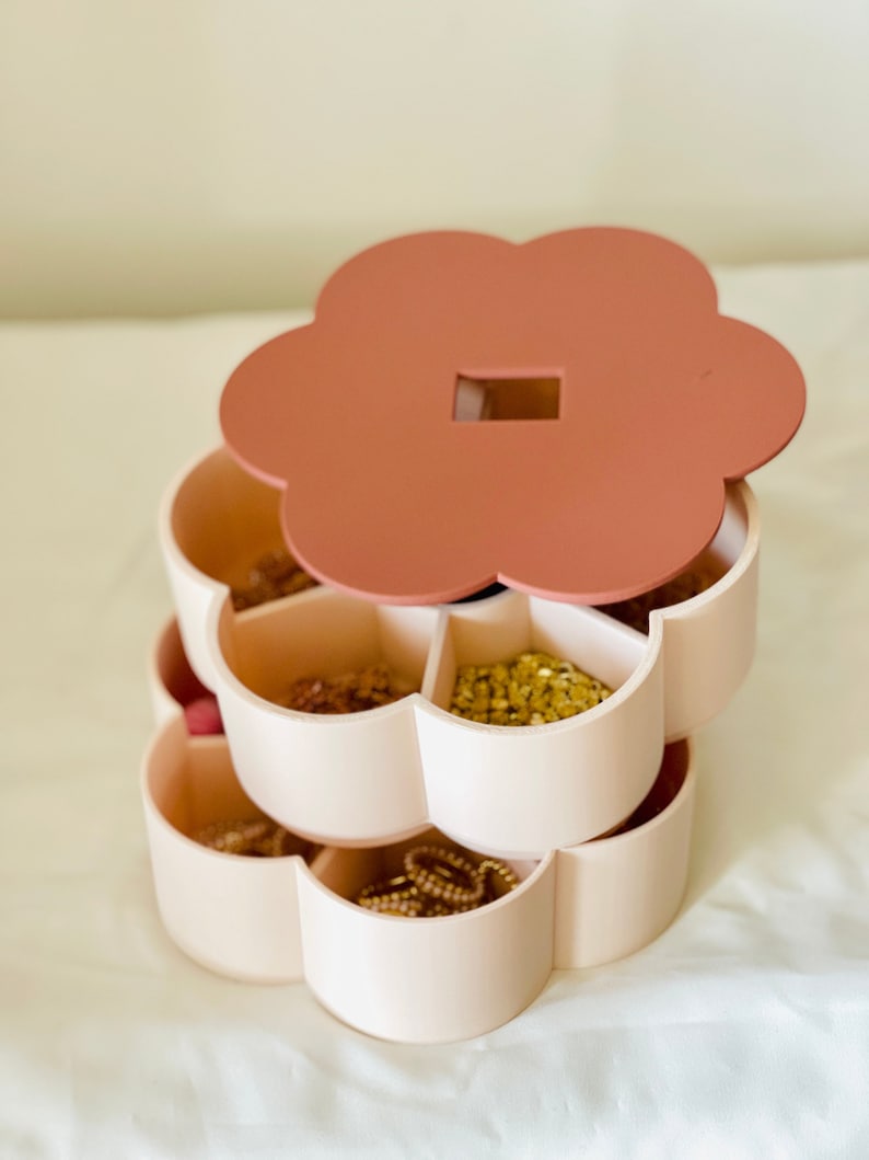 K&ouml;nnte beinhalten: Ein dreistufiger Schmuck-Organizer in Blumenform. Die Oberseite und der Deckel sind in einem ged&auml;mpften Rosaton gehalten, w&auml;hrend die F&auml;cher hellbeige sind. Die F&auml;cher sind mit verschiedenen Schmuckst&uuml;cken gef&uuml;llt, darunter Halsketten und Ohrringe.