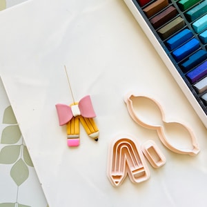 Puede incluir: Una superficie blanca con un lazo rosa, un lápiz amarillo y dos cortadores de galletas de plástico rosa en forma de lápiz y arcoíris. Los cortadores de galletas son para hacer decoraciones de arcilla o fondant.