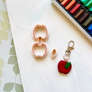 May include: A set of pink plastic apple-shaped cookie cutters, a small leaf-shaped cutter, and a red felt apple keyring with a green felt leaf.