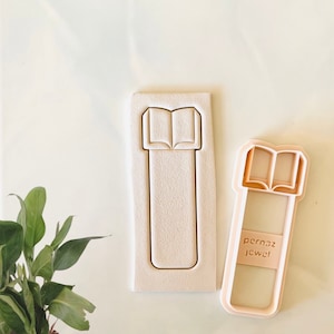 May include: A pink plastic cookie cutter shaped like a book with the text "pernaz jewel!" on the side. The cookie cutter is next to a white silicone mould of the same shape.