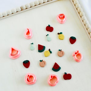 May include: A collection of pink and red fruit-shaped cookie cutters and fondant decorations. The cutters include a watermelon, apple, pear, peach, and strawberry. The fondant decorations include a watermelon, apple, pear, peach, and strawberry.