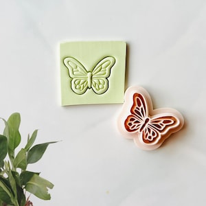 May include: A set of two butterfly-shaped cookie cutters. One cutter is green and the other is pink with a brown butterfly design.