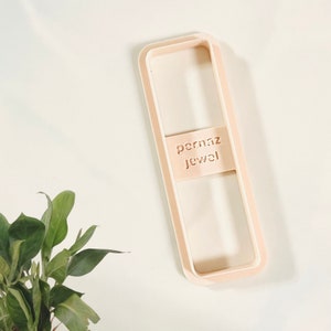 May include: A light brown plastic cookie cutter in the shape of a rectangle with rounded corners. The cookie cutter has the words "pernaz Jewel" cut out on the front.