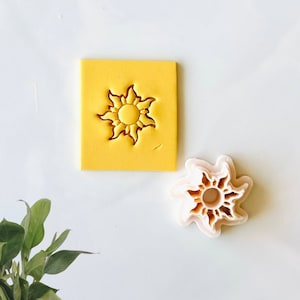 Op de afbeelding: Een gele vierkante koekjesstempel met een zonnesterontwerp en een aparte zonnestervormige koekjesuitsteker. Het zonnesterontwerp is in de gele stempel gesneden. De koekjesuitsteker is lichtroze.