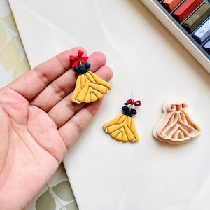 May include: Handmade polymer clay earrings in the shape of a yellow dress with a blue top and red bow. One earring is held in a hand, with another earring and a dress-shaped clay cutter visible.