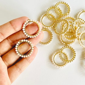 Puede incluir: Primer plano de una mano sosteniendo dos aros de metal dorado con un círculo de perlas blancas alrededor del borde. Hay varios aros más del mismo tipo esparcidos sobre una superficie blanca.