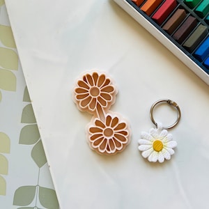 May include: Two plastic daisy-shaped cookie cutters and a white daisy-shaped keyring with a yellow centre. The keyring is on a silver key ring. The cookie cutters and keyring are on a white surface.