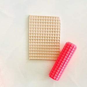 May include: A pink rolling pin with a square pattern and a beige square textured sheet.