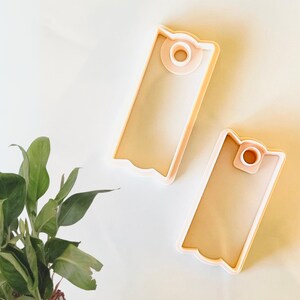 May include: Two plastic cookie cutters shaped like gift tags. The cutters are a light peach color and have a hole in the top for hanging.