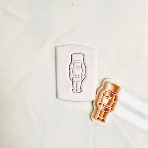 Puede incluir: Un cortador de galletas de plástico blanco con forma de cascanueces. El cortador se muestra en dos vistas, una con el diseño hacia arriba y la otra con el diseño hacia abajo.