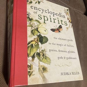 May include: A hardcover book titled Encyclopedia of Spirits by Judika Illes. The cover features a cream background with illustrations of fairies, butterflies, and foliage. It has a bright red spine and text detailing it as a guide to magic.