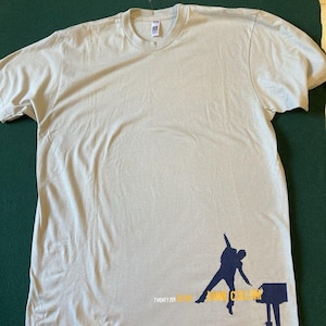 May include: A light beige t-shirt featuring a navy blue silhouette of a person playing a piano. The text "TWENTY ZER" and "JAMIE CULLUM" is printed in navy blue and yellow. The shirt is made of a soft material.