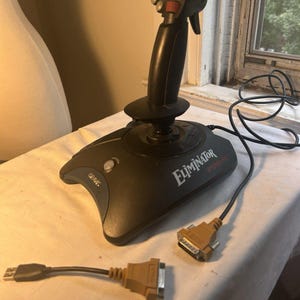 May include: A black Gravis Eliminator joystick with a vertical handle featuring multiple buttons and a red trigger. The base displays the text Eliminator and Gravis. It includes a black cable with a beige connector and a separate beige adapter cable.