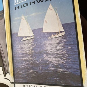 May include: A vintage Mississippi Highways official highway map featuring a blue-toned illustration of two white sailboats on the water. The sailboats are marked with the numbers 10 and 12. The text reads Mississippi Highways and Official Highway Map.