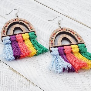 Op de afbeelding: Regenboog-oorbellen met houten regenboog-uitsnijdingen en kleurrijke kwastjes. De regenboog-uitsnijdingen hebben een houtnerf en zijn versierd met kleine, kleurrijke stippen. De kwastjes zijn gemaakt van garen in tinten blauw, paars, roze, geel en groen.