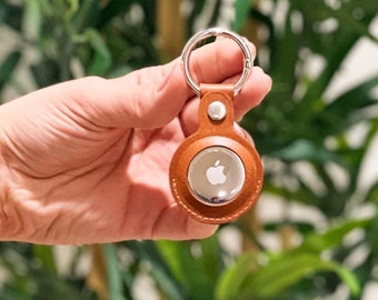 Op de afbeelding: Een bruine leren sleutelhanger met een zilveren metalen cirkel met het Apple-logo. De sleutelhanger wordt in een hand gehouden tegen een groene, bladerrijke achtergrond.