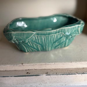 May include: A rectangular ceramic planter with a glossy teal glaze. The exterior features a textured, molded relief pattern resembling overlapping leaves or petals. The rim is uneven and organic in shape, with a deep, hollow center.