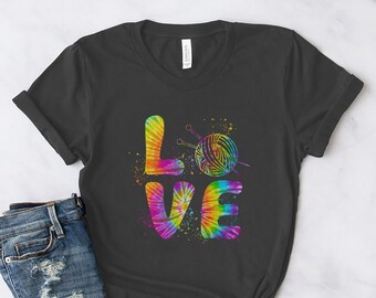 Puede incluir: Una camiseta negra con un diseño tie-dye que deletrea "LOVE" con una bola de lana y agujas de tejer en el centro.