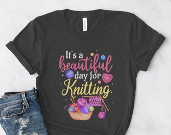 May include: A dark gray t-shirt with the text "It's a beautiful day for Knitting" in pink and white lettering. The shirt features a colorful illustration of knitting needles, yarn, and buttons.