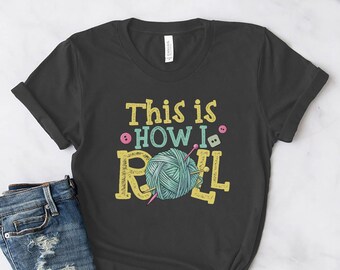Puede incluir: Una camiseta gris oscuro con el texto "This is how I roll" en letras amarillas. Una bola de hilo con agujas de tejer está en el centro del texto.