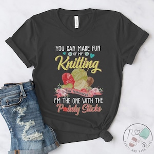 Op de afbeelding: Een donkergrijs T-shirt met de tekst "You can make fun of my knitting but remember... I'm the one with the painty sticks". De tekst wordt omringd door kleurrijke bloemen en wolbollen.