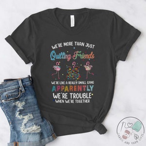 May include: A dark gray t-shirt with a colorful graphic design featuring flamingos and a patchwork quilt. The text on the shirt reads "We're more than just Quilting Friends. We're like a really small gang. Apparently we're trouble. When we're together."