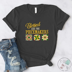 May include: A dark gray t-shirt with the text "Blessed are the Peacemakers" in gold lettering. Three colorful quilt squares are below the text.