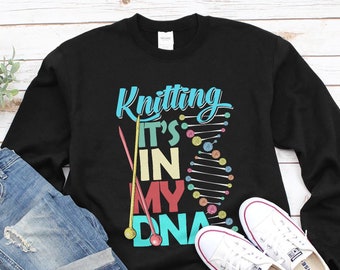 May include: Black sweatshirt with the text "Knitting It's In My DNA" in colorful letters. The text is surrounded by knitting needles and yarn balls. The sweatshirt is laid out on a white wooden surface with a pair of white sneakers.