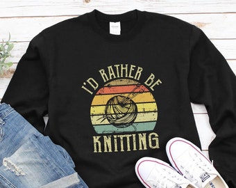 Puede incluir: Sudadera negra con un gráfico de estilo vintage que dice "I'd Rather Be Knitting" con un ovillo de lana y agujas de tejer en un diseño de puesta de sol.