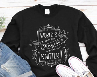 Puede incluir: Sudadera negra con un diseño gráfico blanco que dice "World's Okayest Knitter" en una fuente de estilo vintage.