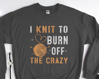 Puede incluir: Una sudadera gris oscuro con el texto "I knit to burn off the crazy" en blanco con una bola de lana naranja estilizada y agujas de tejer.