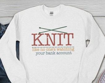 May include: White long-sleeve sweatshirt with a graphic design of crossed knitting needles and the text "KNIT like no one's watching your bank account".