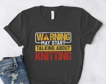 Puede incluir: Una camiseta gris oscura con una señal de advertencia amarilla y roja que dice "Advertencia - Puede empezar a hablar de tejer".
